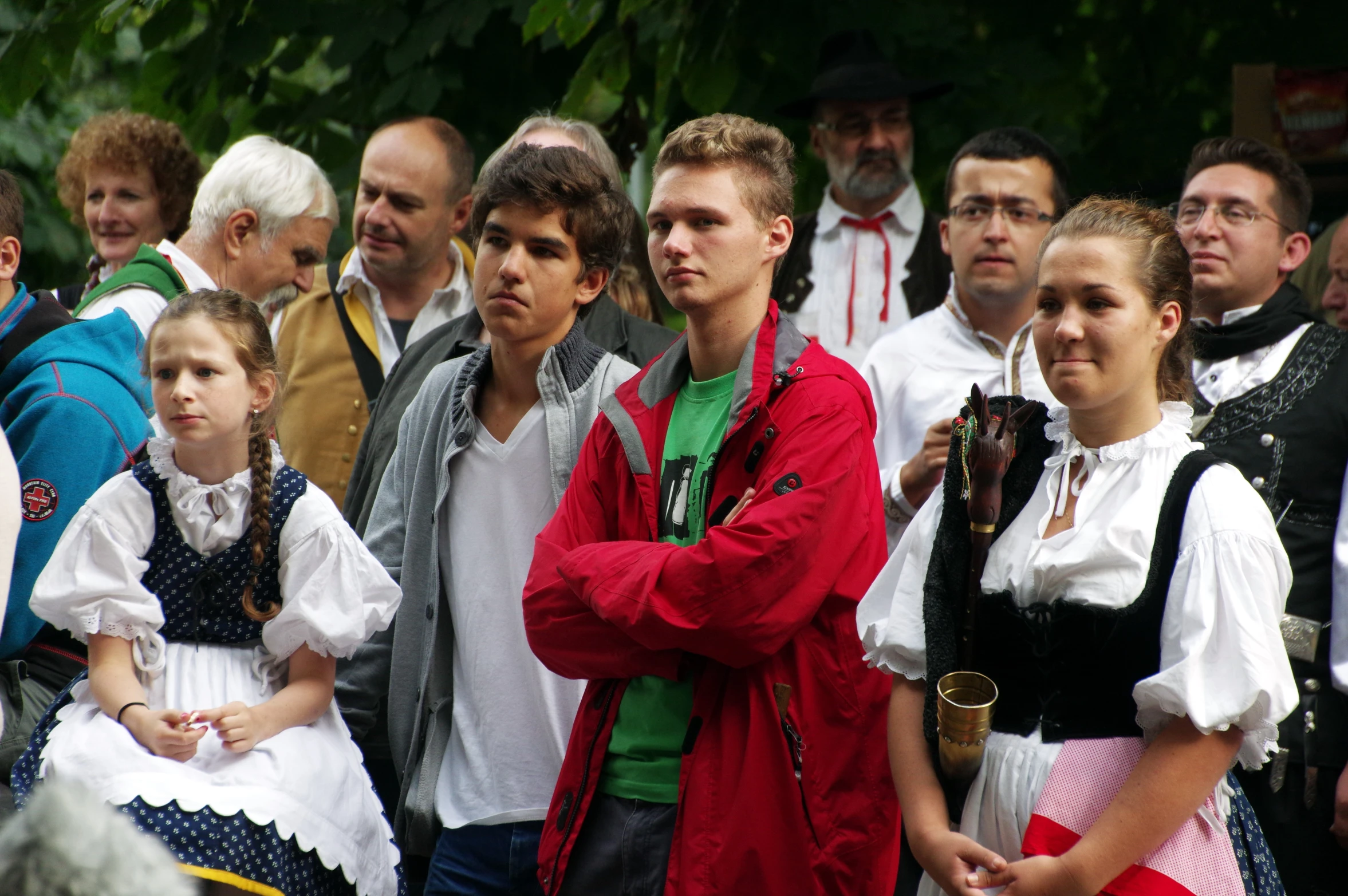 people watching a parade on the street while the person in the white blouse and woman with her arms crossed is talking to someone with a red jacket