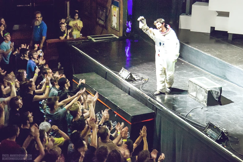 man in white standing on stage with hands raised with crowd in foreground