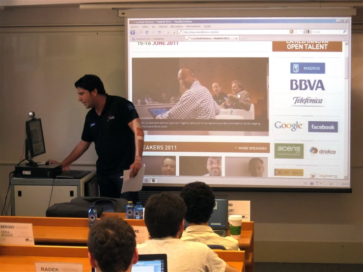 a man stands in front of a class room in front of a projector screen and speaks