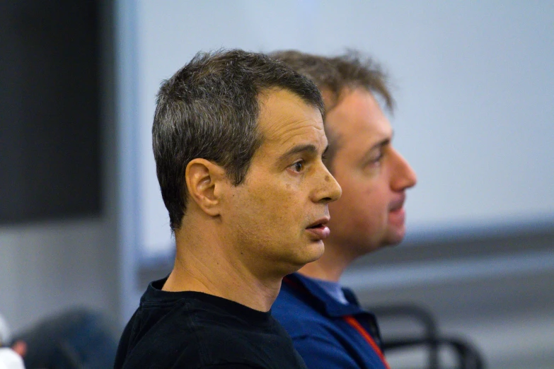 two men look on from behind a black bench