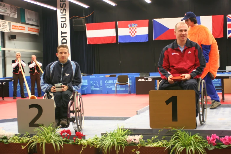 two men sitting in a wheel chair and standing around with one man on top of the other