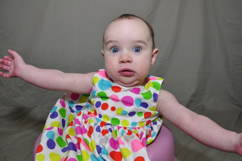 a baby with big blue eyes and colorful polka dot dress holding her arms out