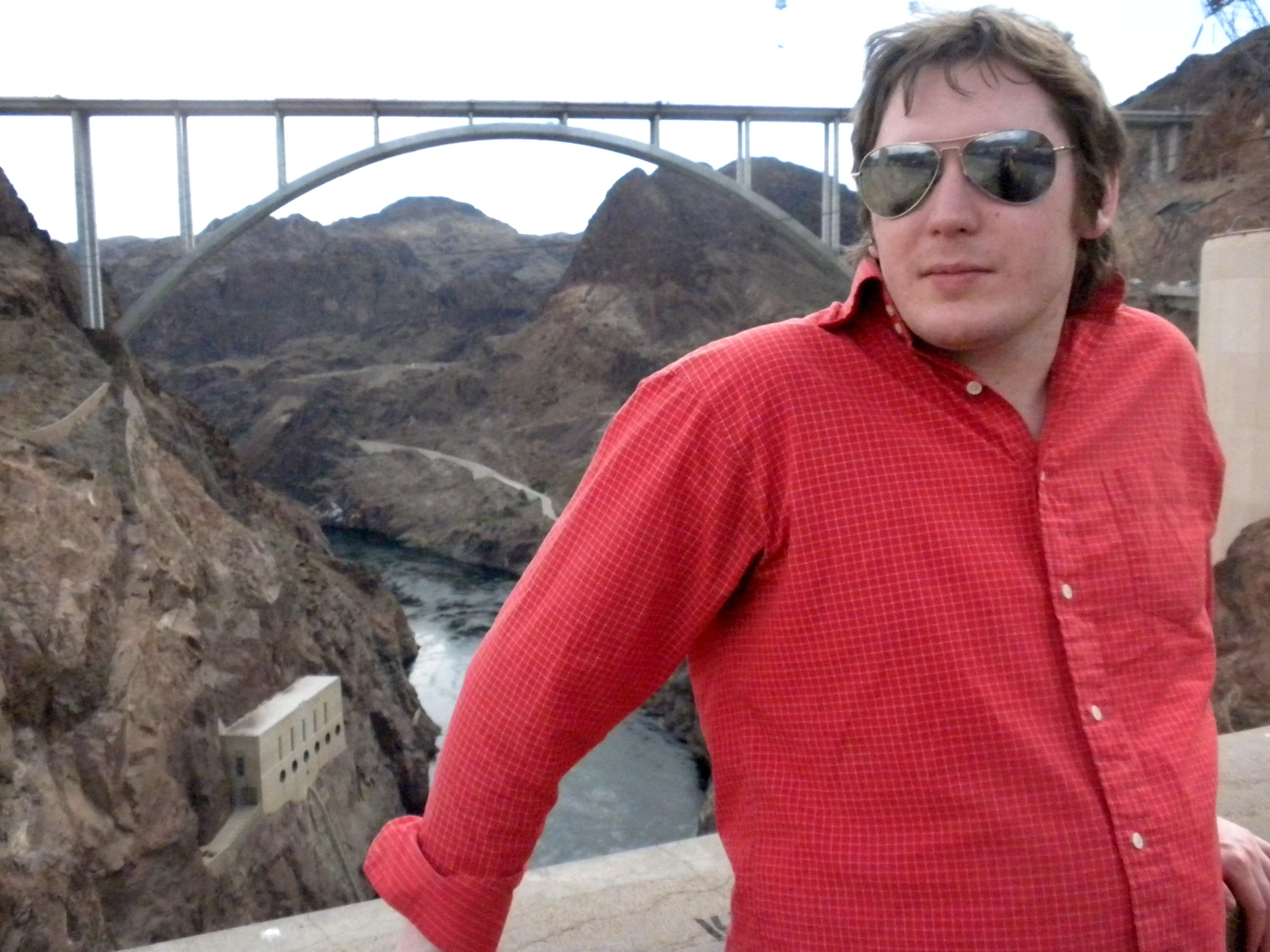 a man wearing sunglasses standing by a bridge with a river below