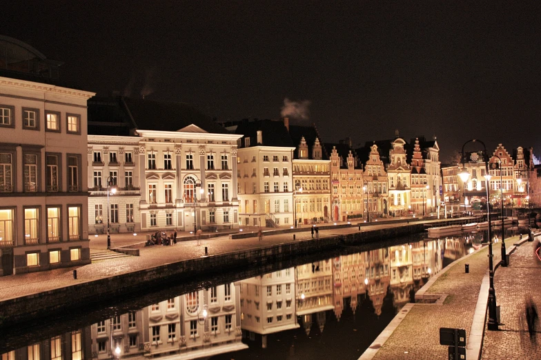 a city night scene with illuminated buildings, reflecting in a canal and people walking on the pavement