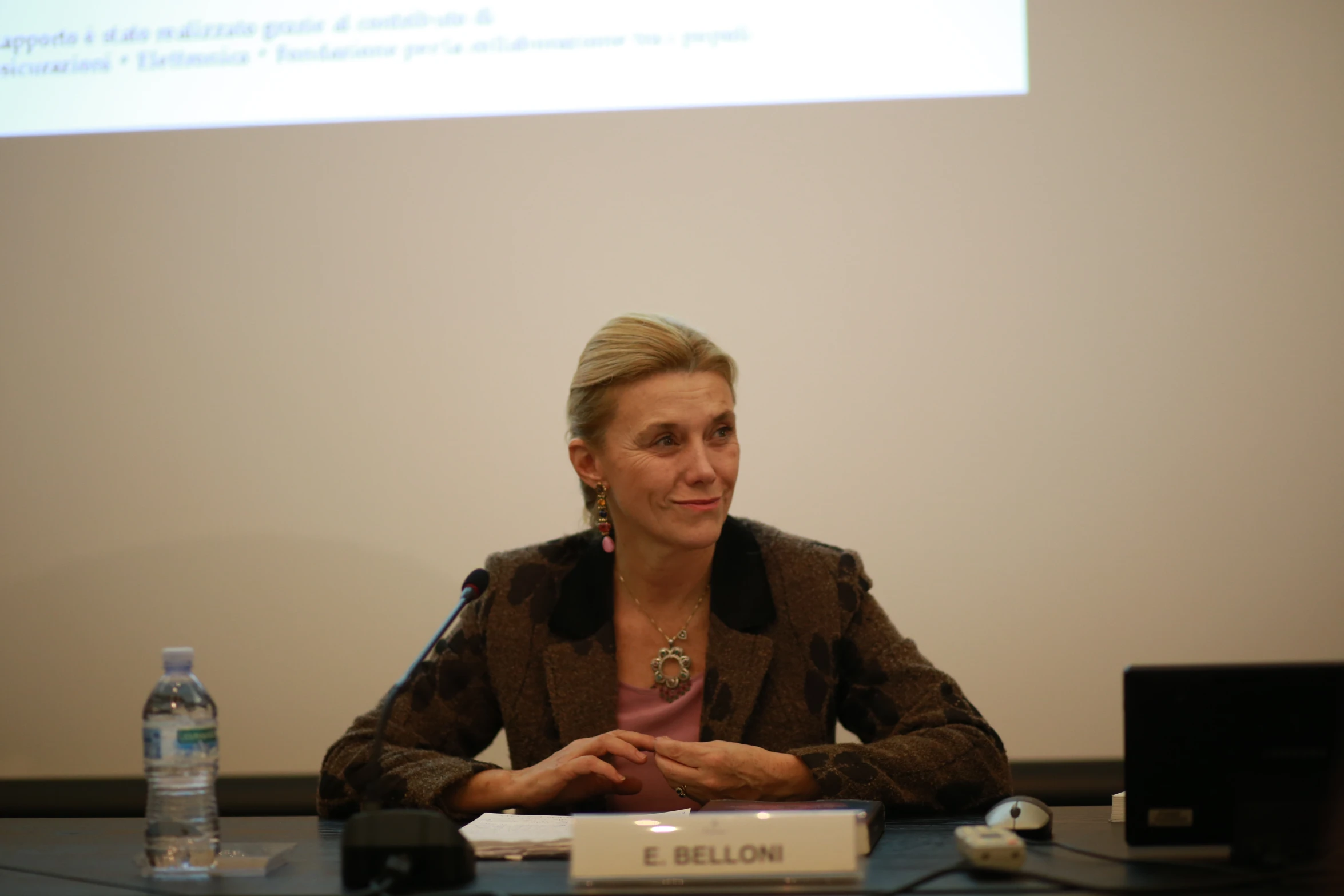 a woman in brown jacket sitting at a table with microphone in front of her