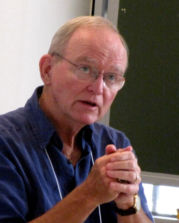 an elderly man wearing glasses, sitting with his hands folded out