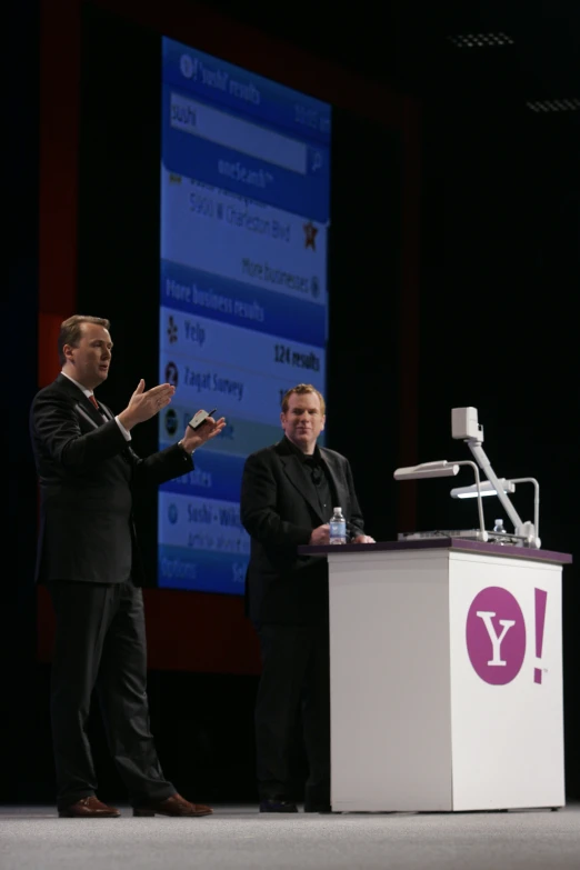 two men standing at podium with a microphone and another man on phone