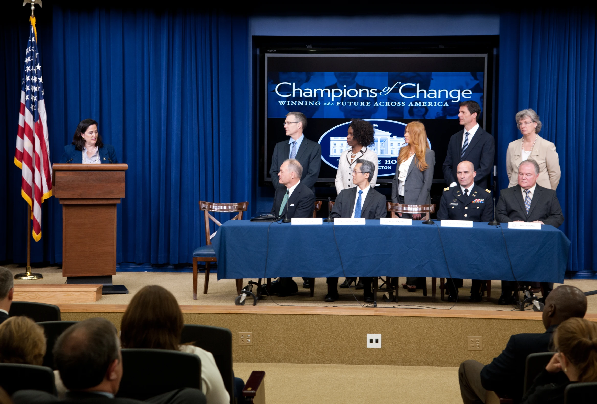 a group of people sitting on a stage and a man standing at a podium