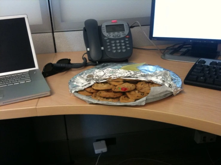 a wooden desk with a bag of cookies on it next to a laptop and telephone