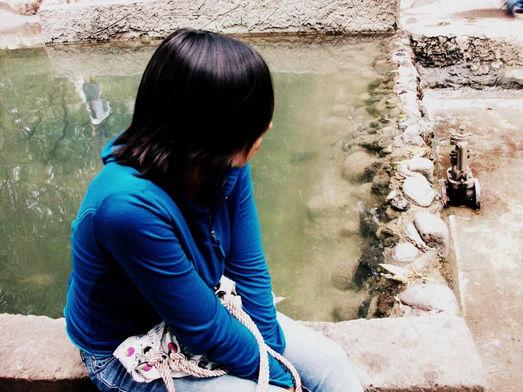 the woman sits near the water, holding her hand near her face