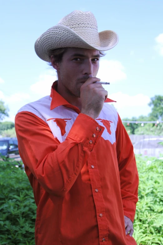 a man wearing an orange shirt and cowboy hat is smoking a cigarette