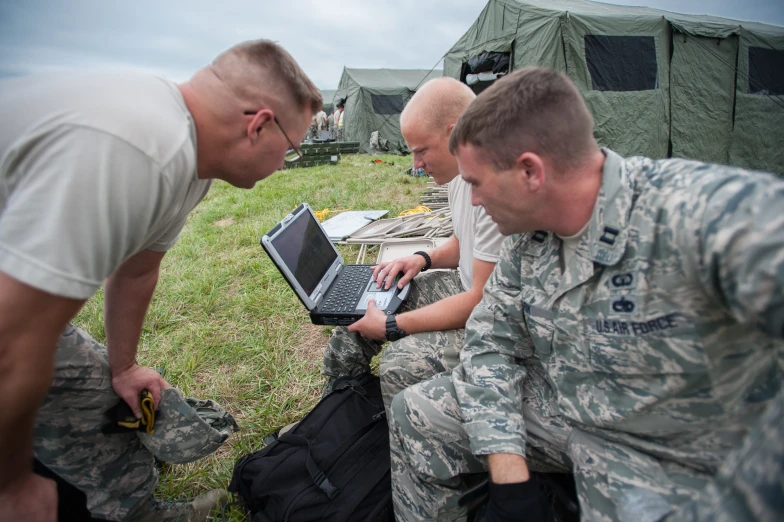 three soldiers sit on the ground while one works on his laptop