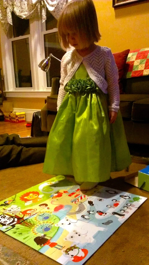 a toddler girl wearing a green dress with an apron standing on the floor in front of a piece of paper