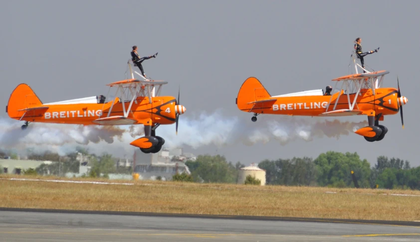 two men in black outfits stand on the wings of an orange plane