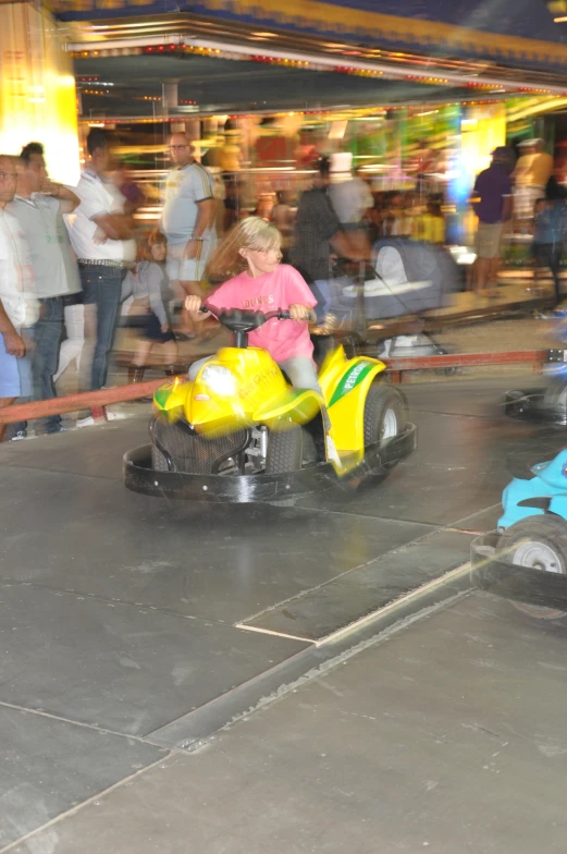 three children in a carnival ride, one wearing pink and yellow, riding on the two wheeled vehicles