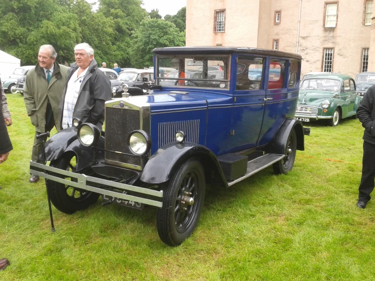 people look at an old - fashioned car parked on the grass in front of some older vehicles