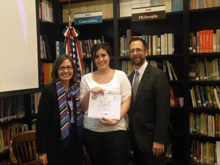 three people standing with a certificate in front of a bookshelf