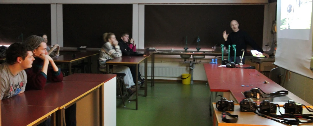 three girls at desks in a classroom with people looking on