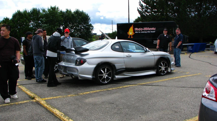 people standing around a silver car in a parking lot