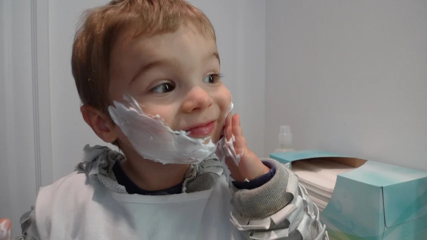 a small boy standing in front of a bathroom mirror with shaving on his face