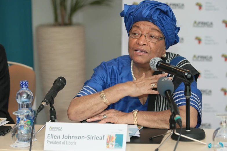 a woman in blue clothing sitting at a table with microphones