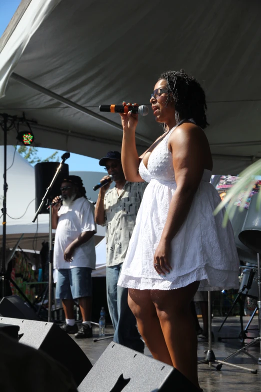 woman in white dress speaking on stage with band playing