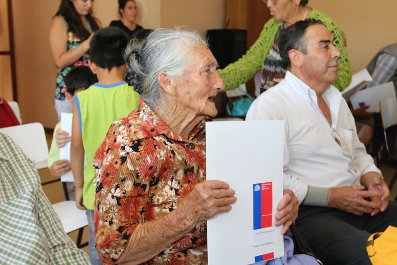 old woman holding up a piece of paper while sitting next to a man in the front of a group of older people