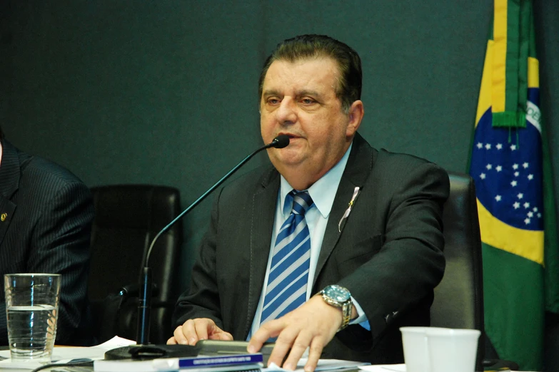 a man with glasses at a meeting and another man sitting at the side of the table