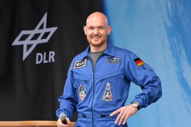 an older man in a blue astronaut suit smiles in front of a black and blue sign