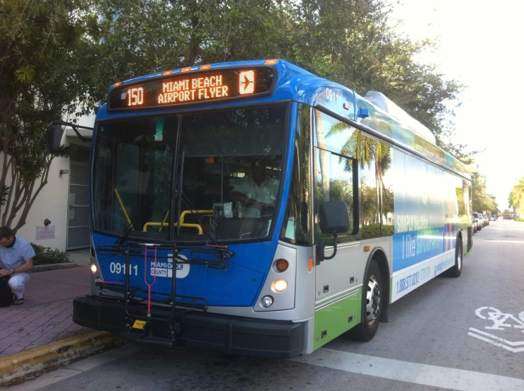 a blue, white, and green bus sits on the side of the road as people sitting in the shade