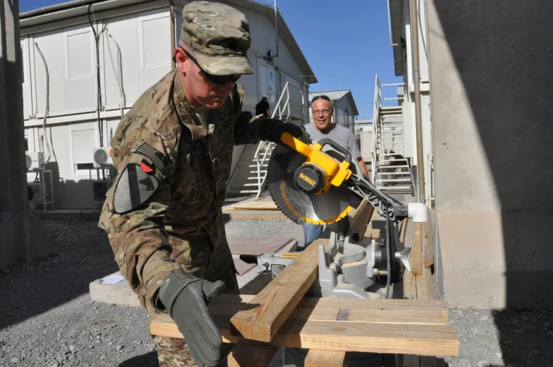 a man using a driller and a man wearing camouflage standing behind him