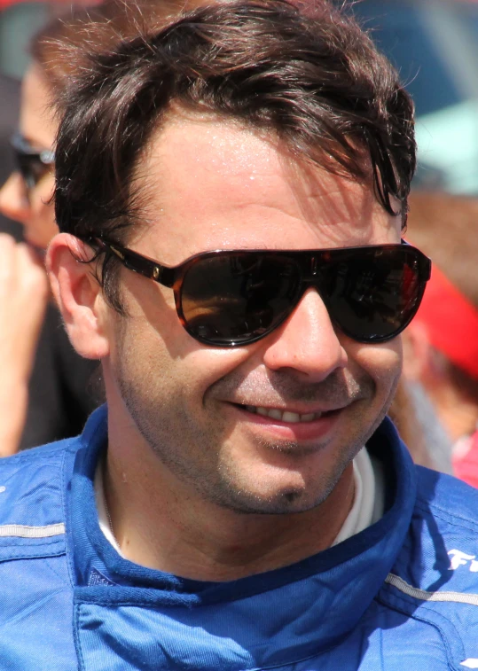 man with sunglasses smiling while wearing a blue racing suit