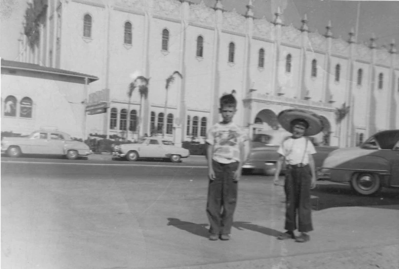 two boys stand in front of a building and one is holding a large black umbrella