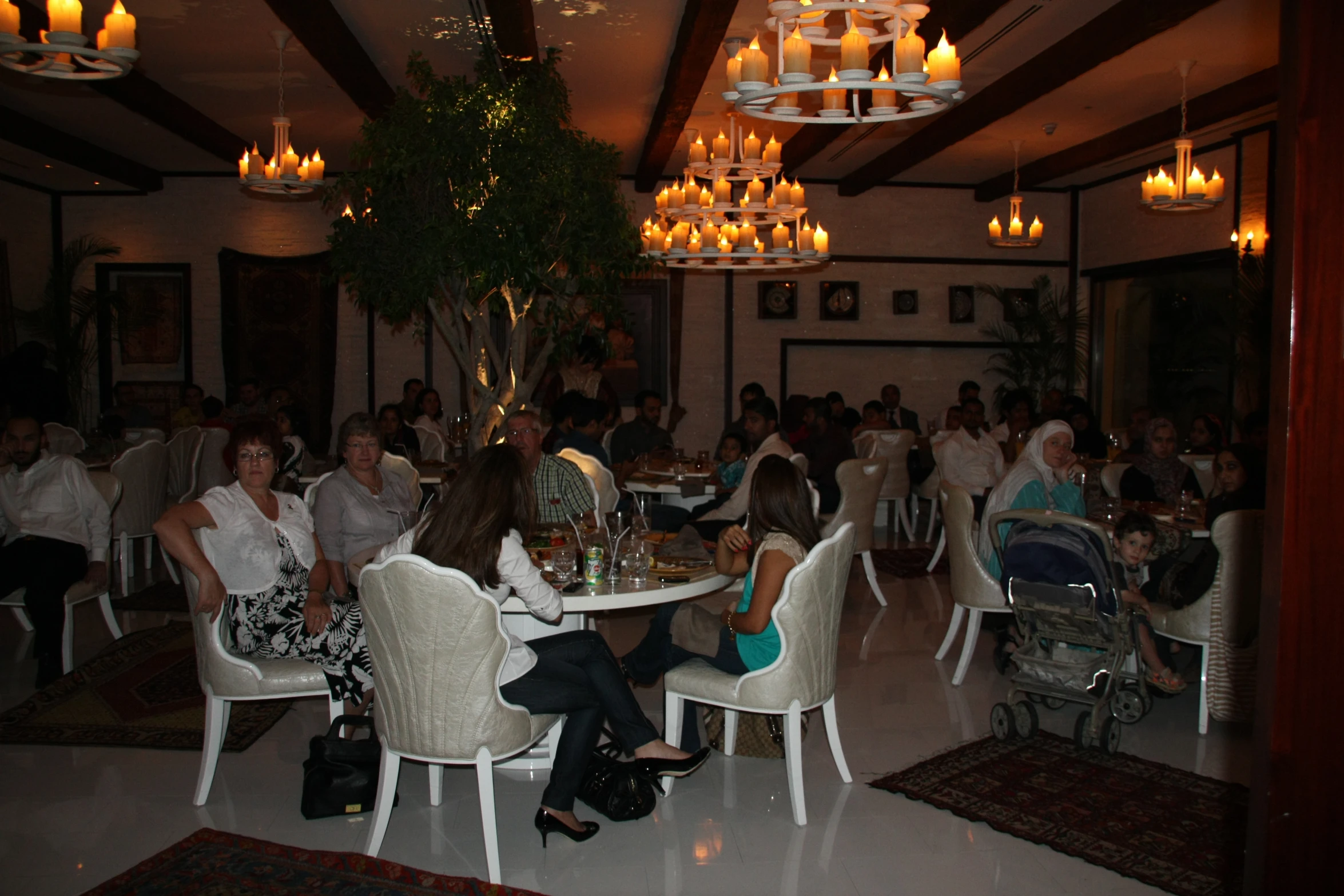 a restaurant with white chairs and chandeliers full of people eating