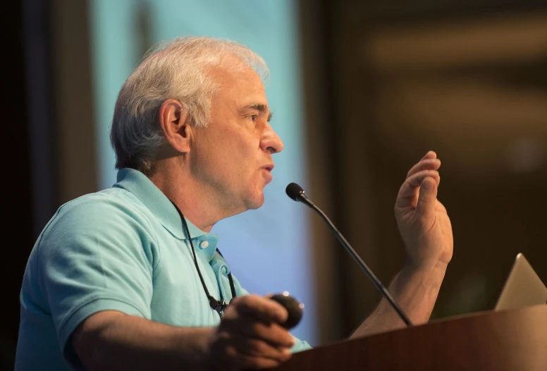 an older man in a light blue shirt gives a speech