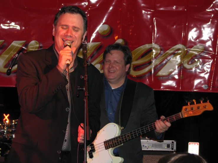 two men laugh while singing into microphones and holding guitars