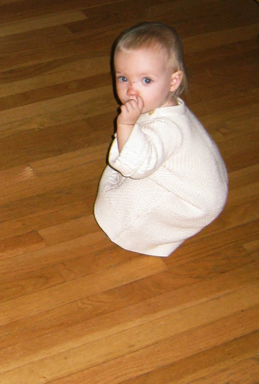 baby in white sweater on hardwood floor chewing with hand