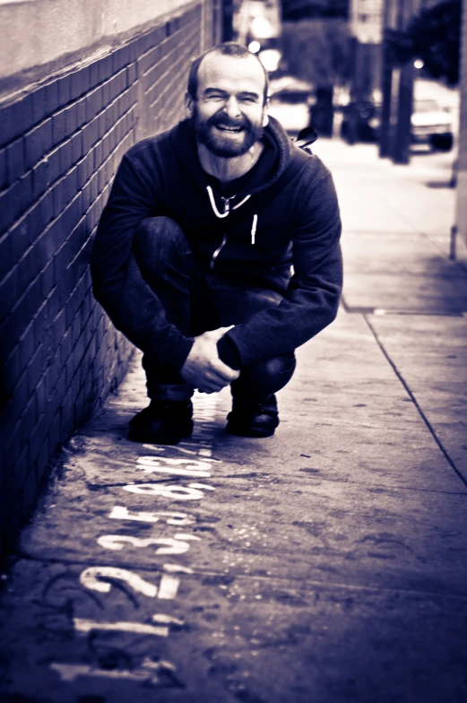 a man crouches on the curb in front of a wall and points at writing on it