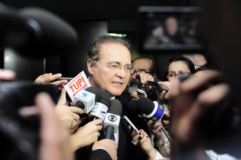 a man in suit and glasses surrounded by reporters holding microphones