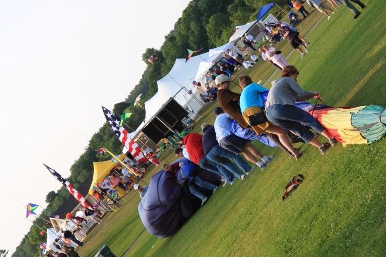 several people sitting and standing on grass near tents and flags