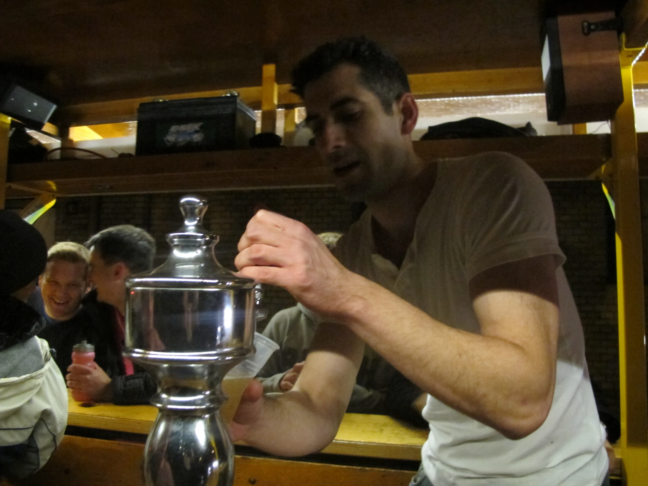 people seated in a small cafe, all with one man pouring coffee on a metal blender