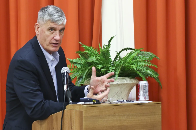 man speaking from podium next to orange curtain