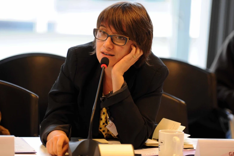 a woman in glasses sits at a table and gestures