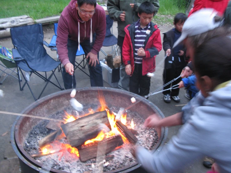 several adults and children are surrounding a bbq grill