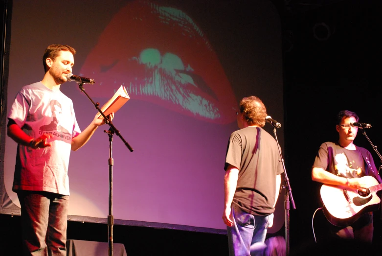 three men are standing on stage, one with an acoustic guitar, the other with a microphone