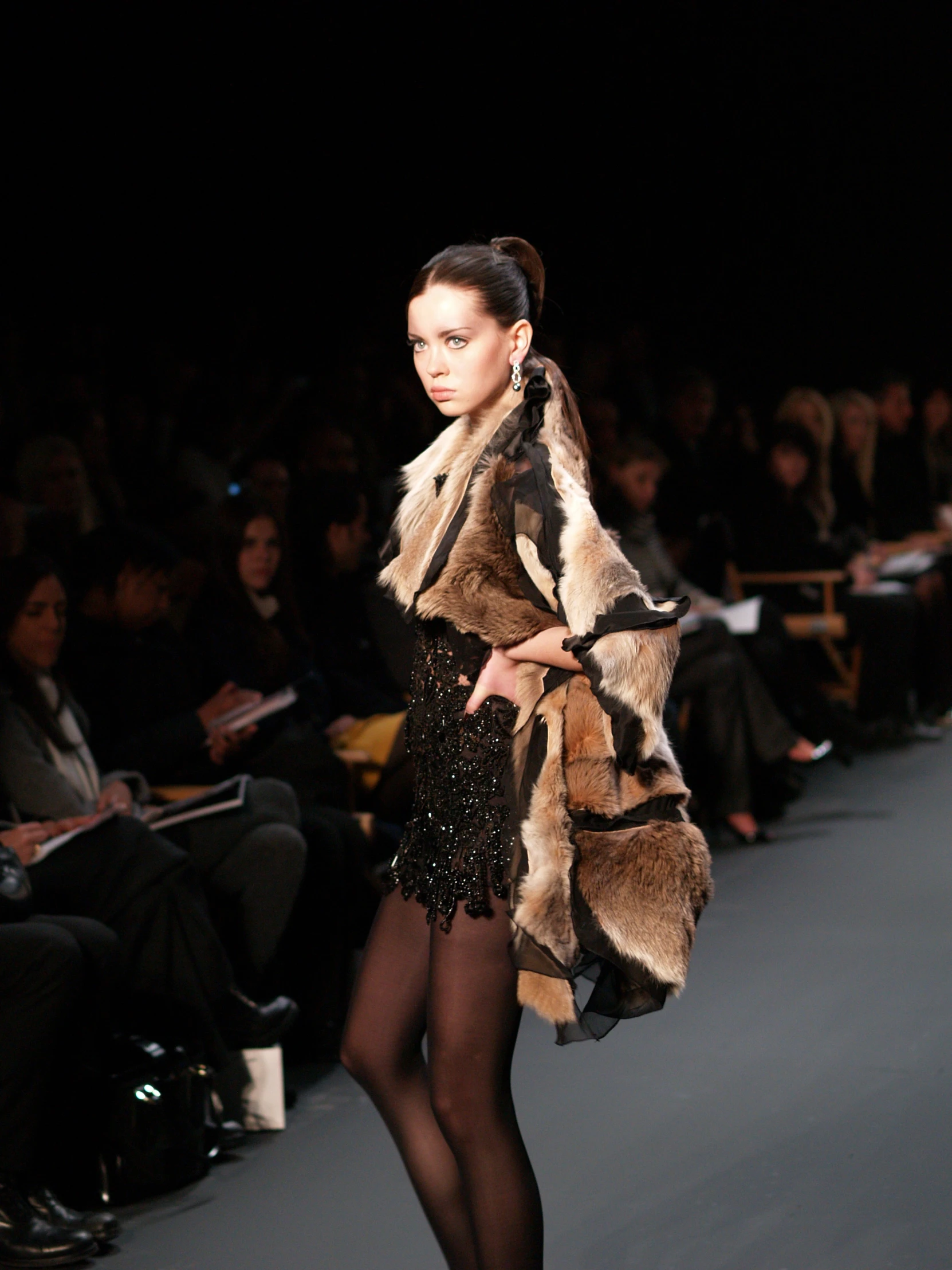 a woman walking down a runway in a black dress and fur coat