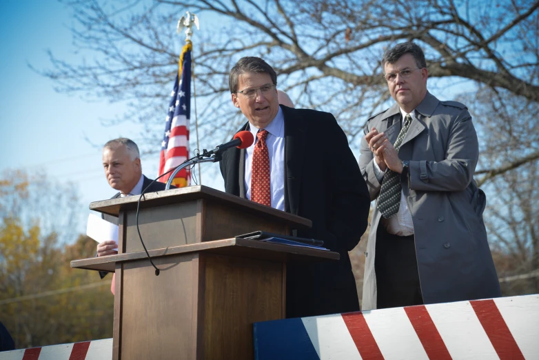 two men standing behind a podium, while one stands behind him