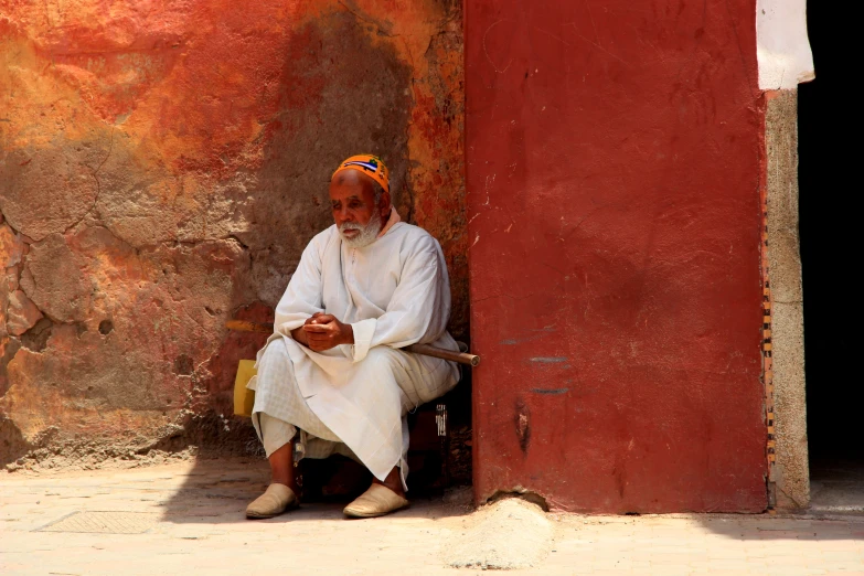 an old man sitting on the side of a building looking at his cell phone