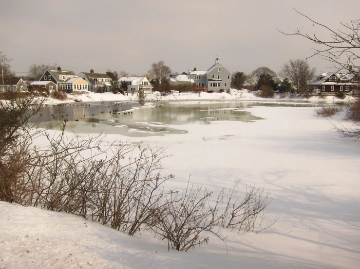 a small lake surrounded by snow with buildings and trees on the shore