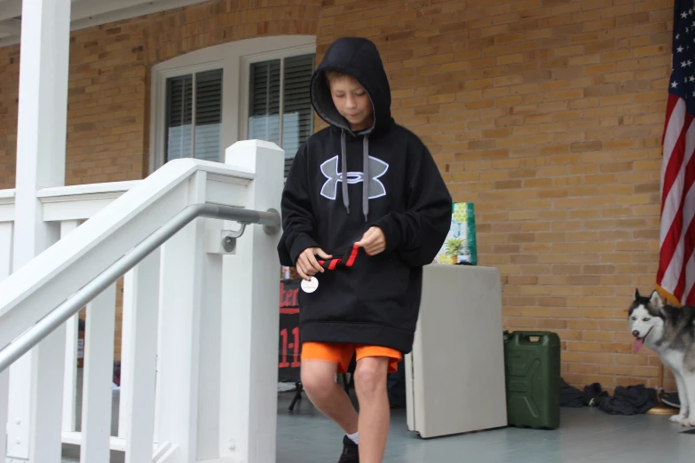 boy in a hoodie walks along a porch holding a tennis racket